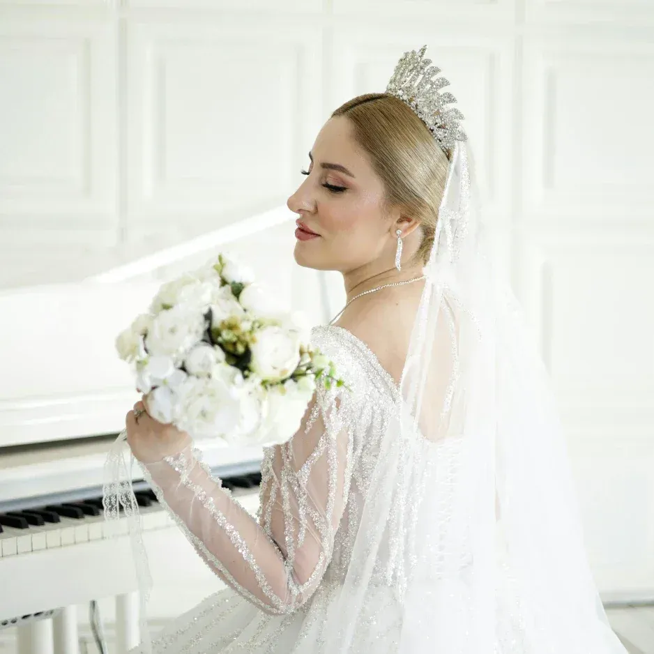 Bride in a white wedding dress holding a bouquet of white flowers, wearing a tiara and veil, standing near a white piano in a bright room.