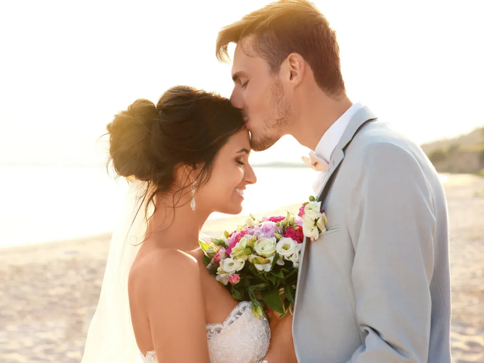 A bride and groom on the beach, with the groom kissing the bride on her forehead while she holds a bouquet of flowers.