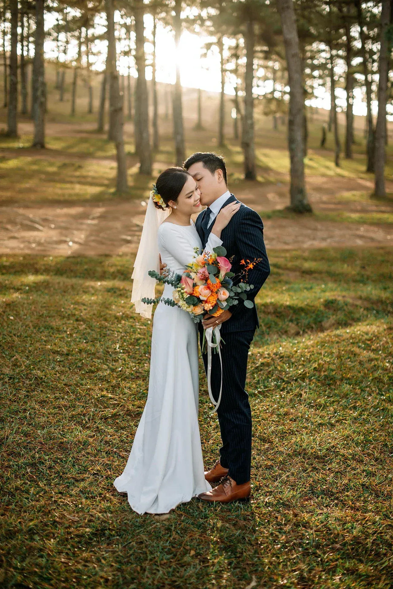 A newlywed couple embracing outdoors in a wooded area during sunset, with the bride holding a vibrant bouquet of flowers.