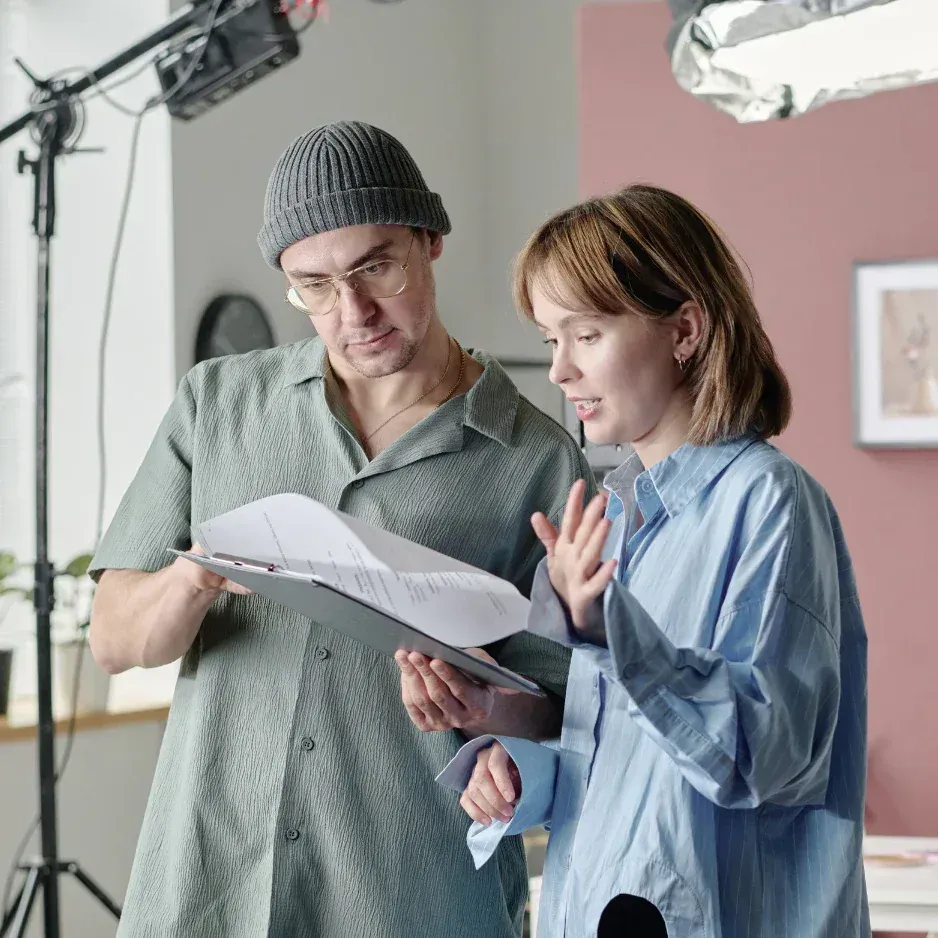 Two individuals reviewing a script or document on a film or video set, with studio lighting and equipment visible in the background.