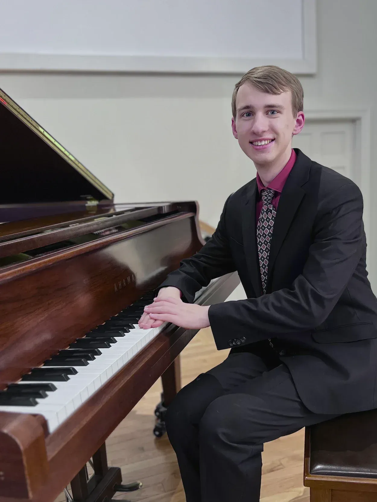 A young man in a black suit and patterned tie playing a brown Yamaha upright piano indoors, smiling at the camera.