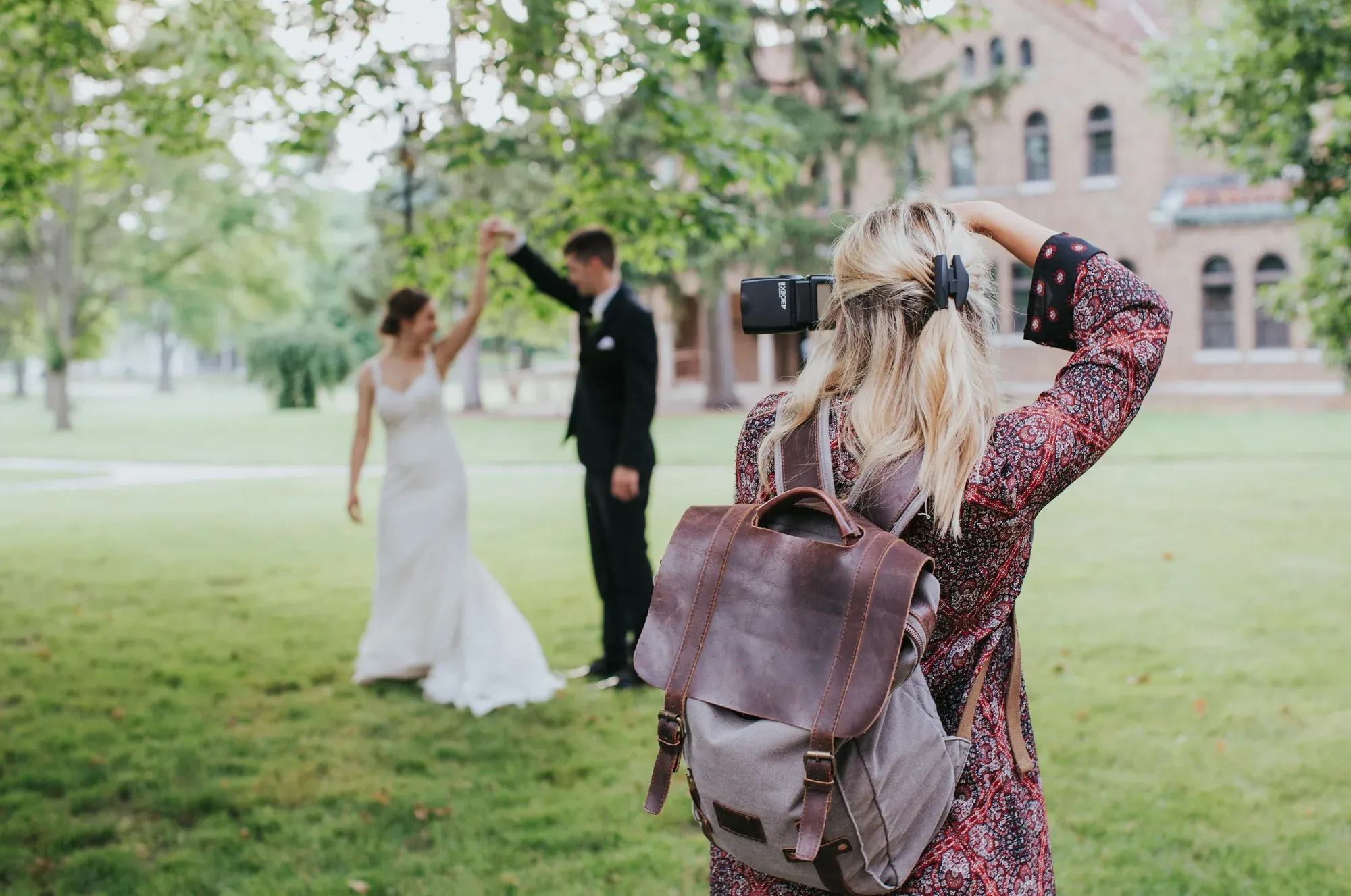 A woman with blond hair and a brown backpack takes a photo of a bride and groom dancing outside on a grassy area with trees and a brick building in the background.