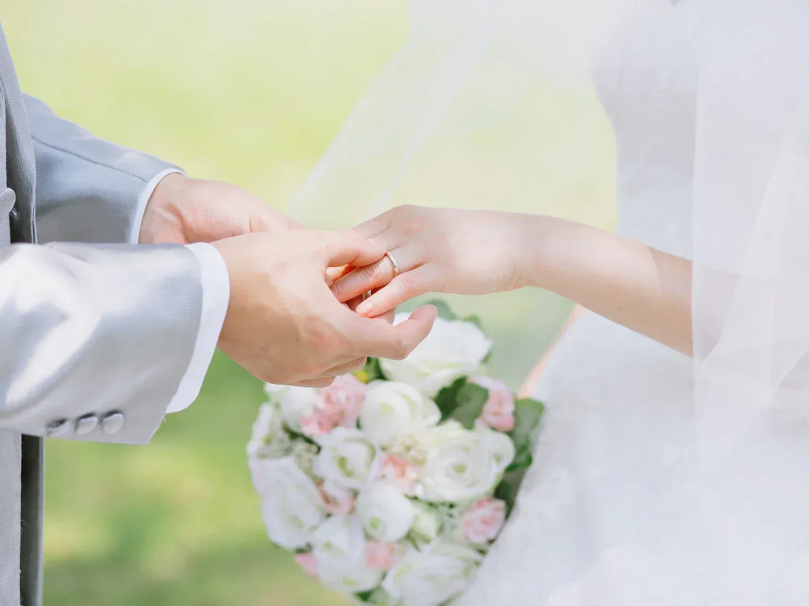A bride and groom hold hands during a wedding ceremony, with the groom placing a ring on the bride’s finger, with a bouquet of white and pink flowers in the background.
