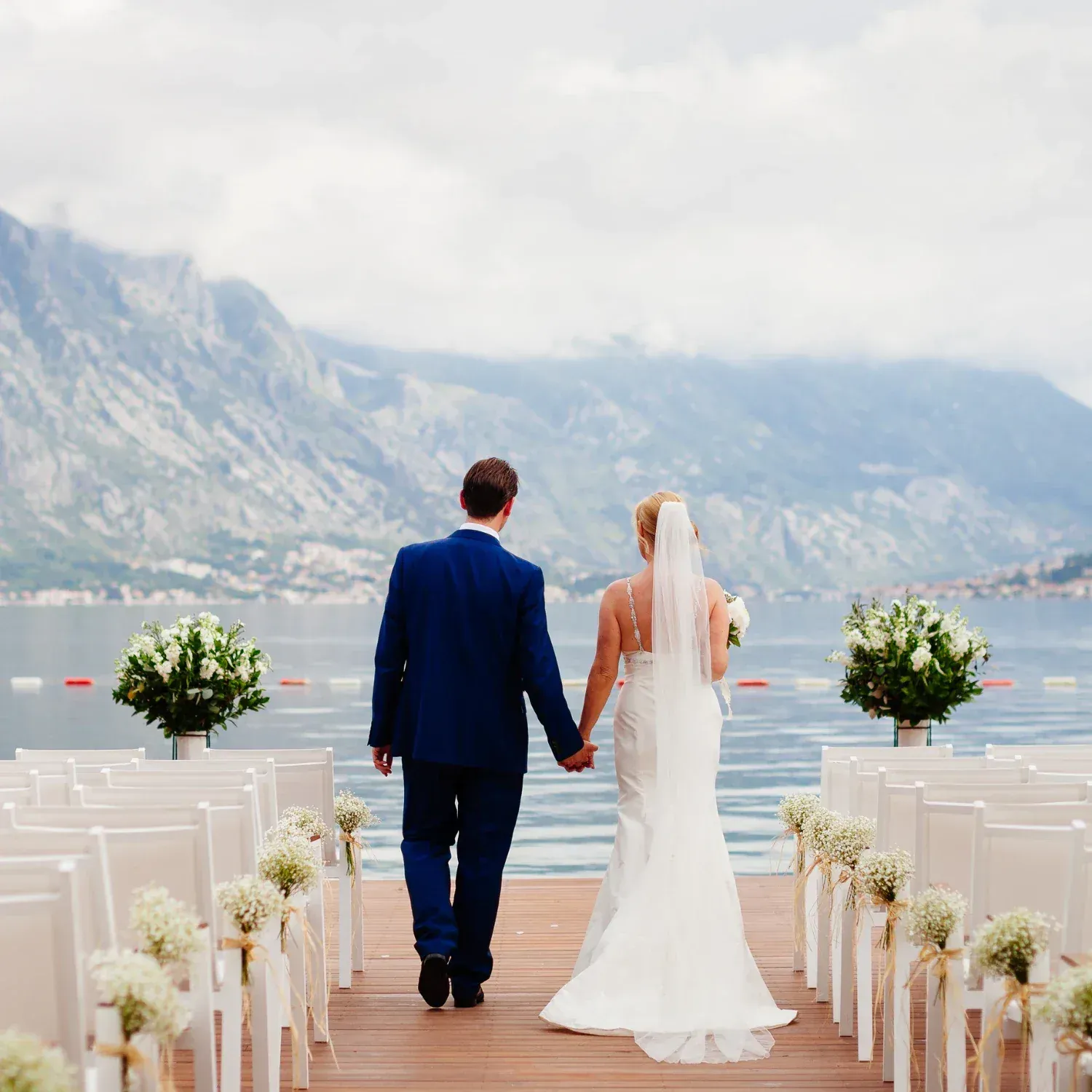 Bride and groom walking hand-in-hand on a dock towards a lake and mountains background during a wedding ceremony.