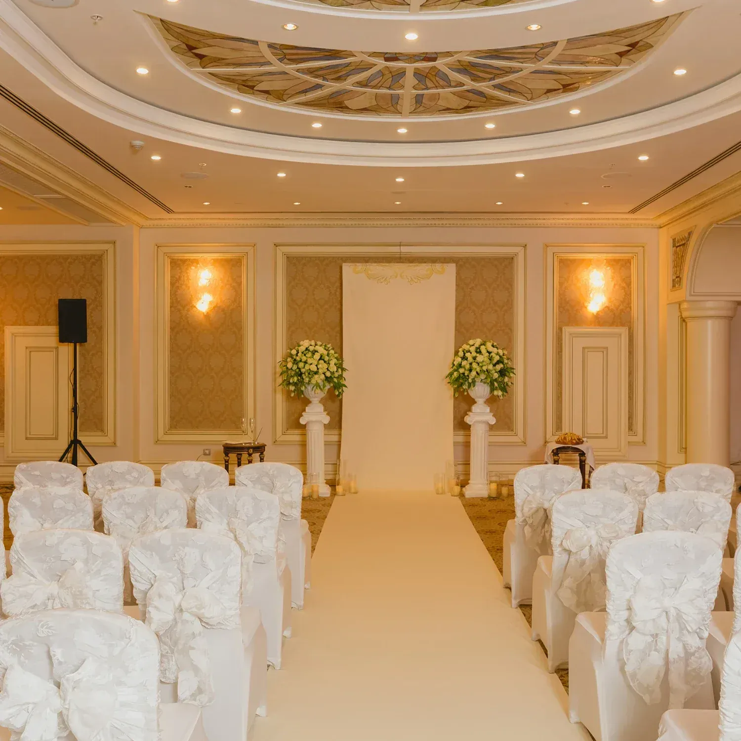 Elegant wedding ceremony setup with white chairs adorned with ribbons, a beige aisle runner, a backdrop of white flowers in vases on pedestals, and a decorated ceiling in a banquet hall.