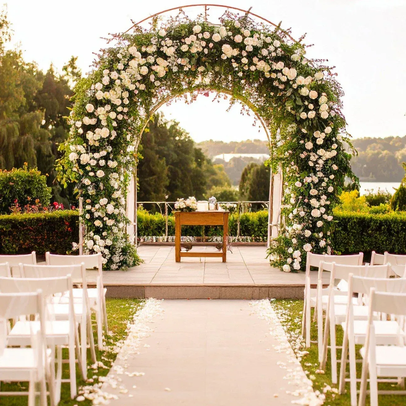 Outdoor wedding ceremony setup with white chairs, flower petals on the aisle, and a floral arch over the altar, overlooking a scenic lake and lush greenery.